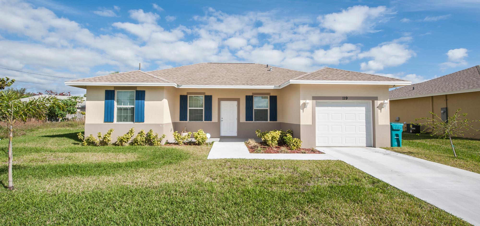 Beige one story house with dark shutters and one car garage and grass lawn